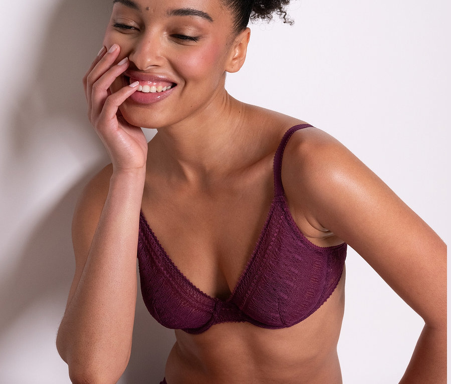 Eine lächelnde Frau sitzt und trägt einen lilafarbenen Passionata Bügel-BH »Leila« und passende Unterteile.