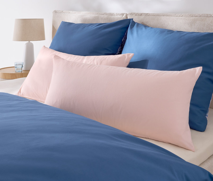 Auf einem Bett liegen zwei blaue und zwei rosa Kissen. Nachttisch mit Lampe und einem Glas Wasser.