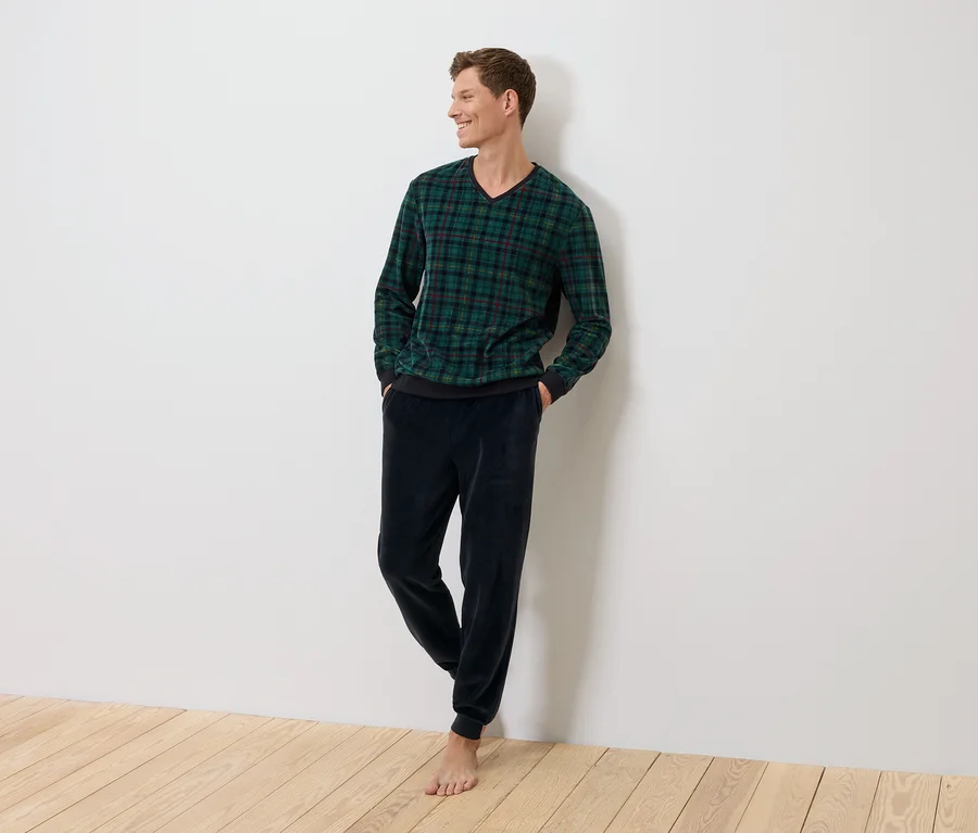 Ein Mann steht an einer Wand, bekleidet mit einem grün karierten Pullover und dunkelblauen Samthosen.