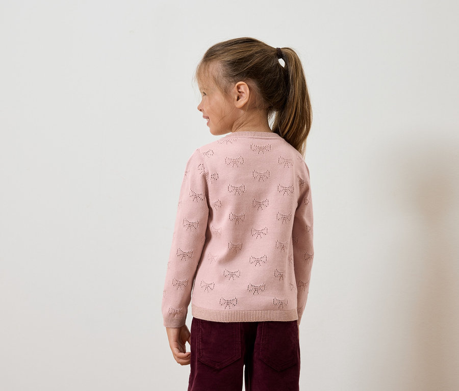 Mädchen mit Zopf trägt rosa Pullover mit Schleifen und weinrote Hose, seitliche Aufnahme.