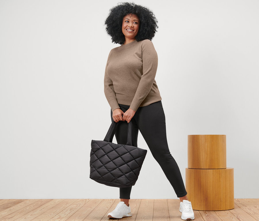 Frau hält eine schwarze Stepptasche und trägt einen beigen melierten Feinstrickpullover, schwarze Leggings und weiße Turnschuhe.
