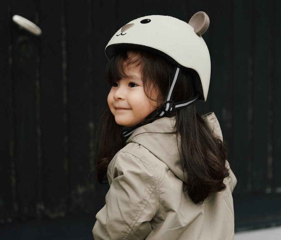 Lächelndes Mädchen mit braunen Haaren, beiger Jacke und HUDORA Kinderhelm, Beary, 48–52 cm auf dem Kopf sitzt auf einem lila HUDORA Laufrad.