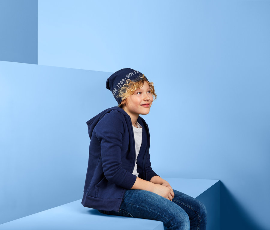 Ein Junge mit blonden Haaren sitzt und trägt eine Wendemütze und eine blaue Kapuzenjacke.