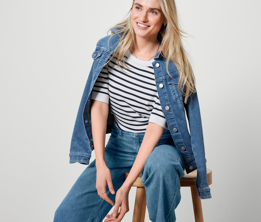 Blonde Frau sitzt auf einem Holzhocker und trägt eine Jeansjacke, Palazzo Jeans und einen gestreiften Feinstrickpullover.