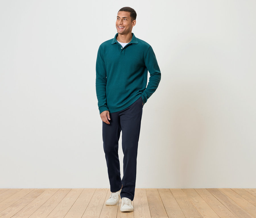 Ein Mann steht auf einem Holzboden und trägt eine dunkelblaue Hose, ein grünes Langarmshirt und weiße Turnschuhe.