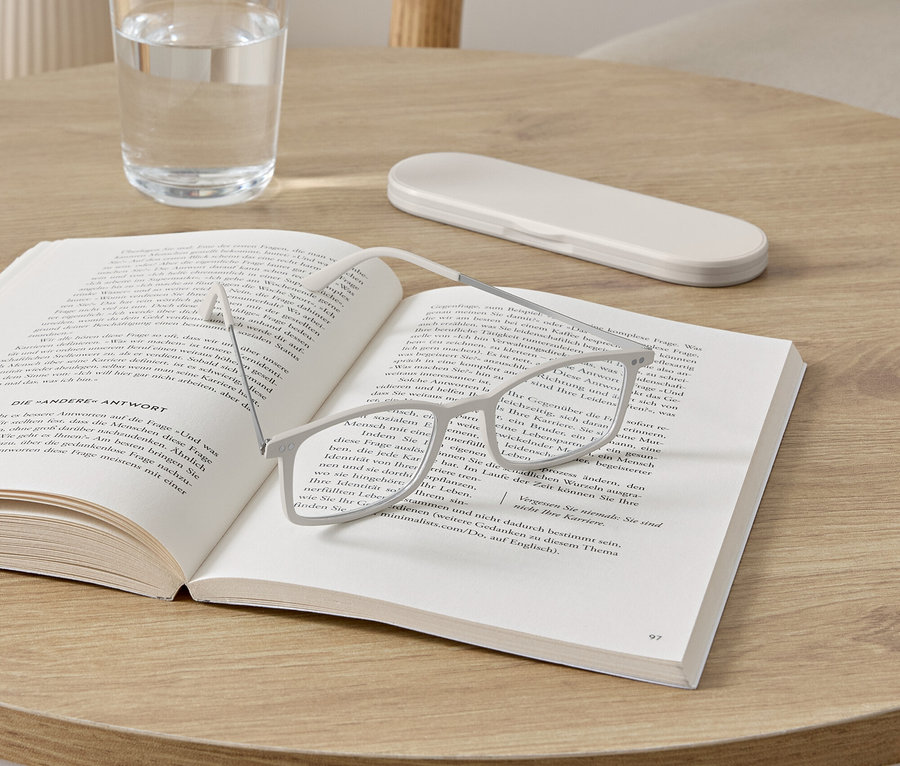 Eine Brille liegt auf einem aufgeschlagenen Buch auf einem runden Tisch. Neben dem Buch stehen ein Glas Wasser und ein Brillenetui.