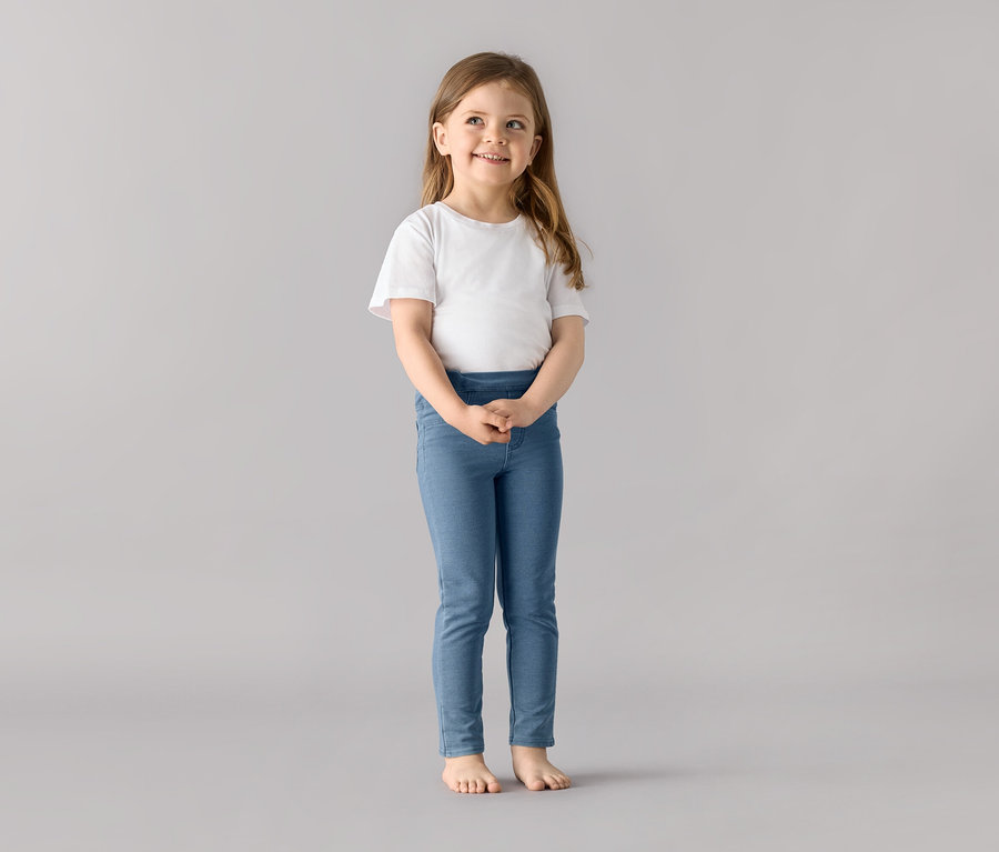 Lächelndes Mädchen steht und trägt ein weißes T-Shirt und blaue 2 Kinder-Jeggings ‒ Fit »Mia«.