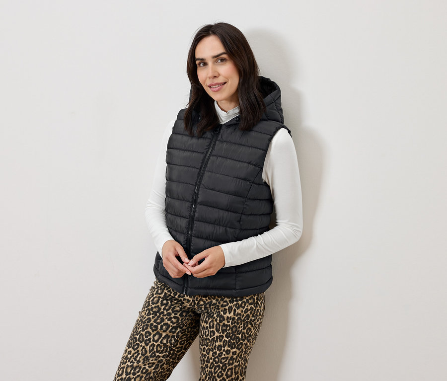 Frau posiert in einer schwarzen Steppweste mit Kapuze, einem weißen Rollkragenpullover und Animal-Print-Hosen.