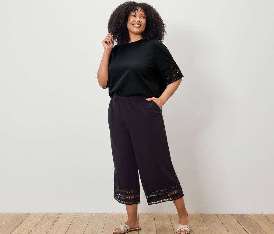 Frau posiert in schwarzem Shirt mit Stickerei und schwarzen Culotte mit Stickerei.