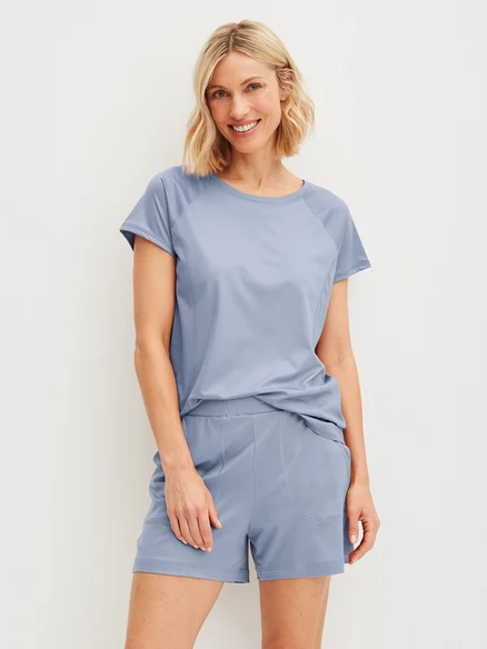 Frau mit blonden Haaren, die ein blaues T-Shirt und Shorts trägt und lächelt.