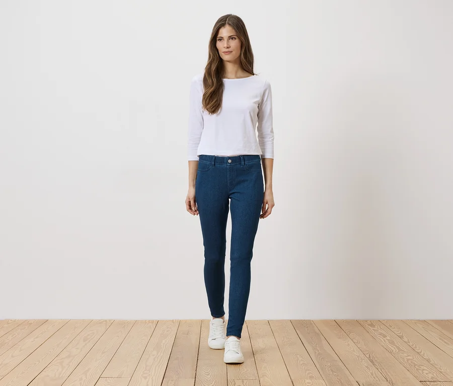 Frau mit weißem Oberteil, blauen Jeans und weißen Schuhen steht auf Holzboden vor einer weißen Wand.