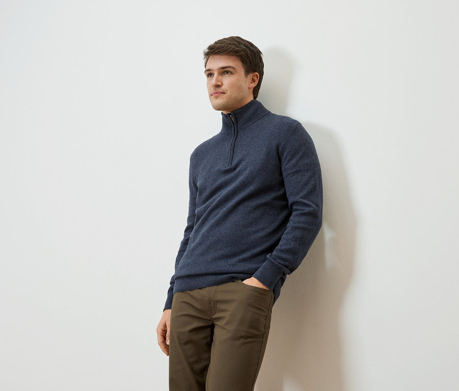 Mann lehnt an einer Wand, trägt einen marineblauen Pullover mit Reißverschluss und khaki-farbene Hosen.