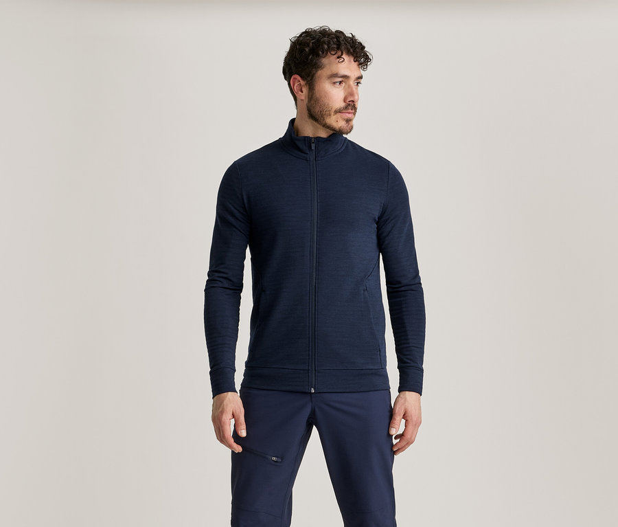 Mann mit dunklen, lockigen Haaren, Bart und Schnurrbart, trägt eine marineblaue Jacke mit Reißverschluss und marineblaue Hose.
