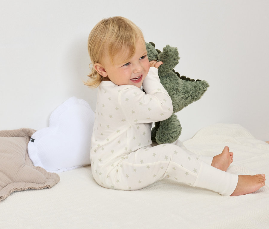 Ein blondes Kind im weißen Sternen-Pyjama sitzt auf einer weißen Decke und umarmt ein grünes Dinosaurier-Stofftier.