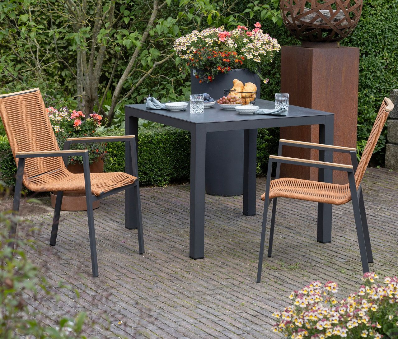 Auf einer Terrasse stehen ein grauer Tisch mit zwei Stühlen mit geflochtenen Sitzen, Gedeck und Speisen.