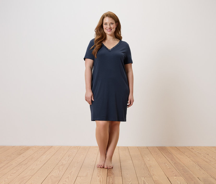 Frau steht barfuß auf einem Holzboden und trägt ein nachtblaues Nachthemd.