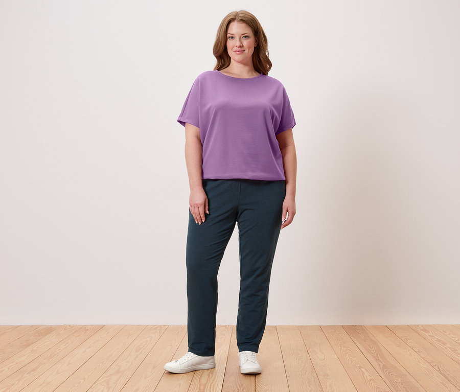 Eine Frau steht vor einer Wand und trägt ein violettes T-Shirt, eine dunkle Hose und weiße Turnschuhe.