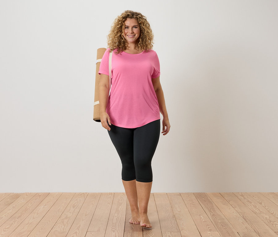 Frau mit lockigem Haar, trägt ein rosa T-Shirt und schwarze Leggings, eine Yogamatte über der Schulter, barfuß auf Holzboden.