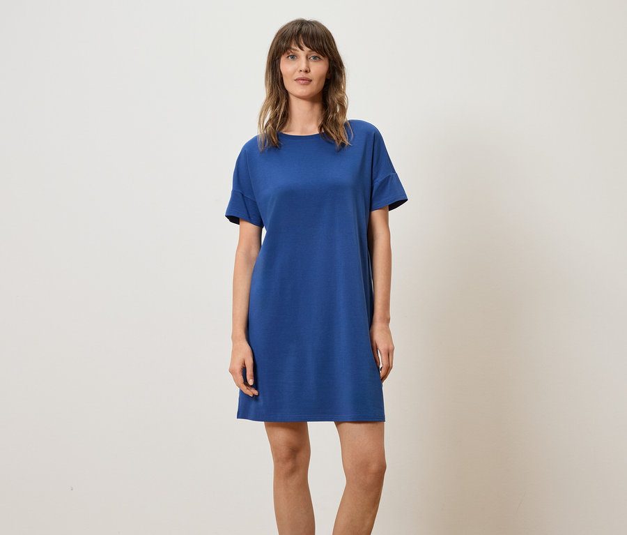 Frau trägt blaues T-Shirt-Kleid vor einem neutralen Hintergrund.