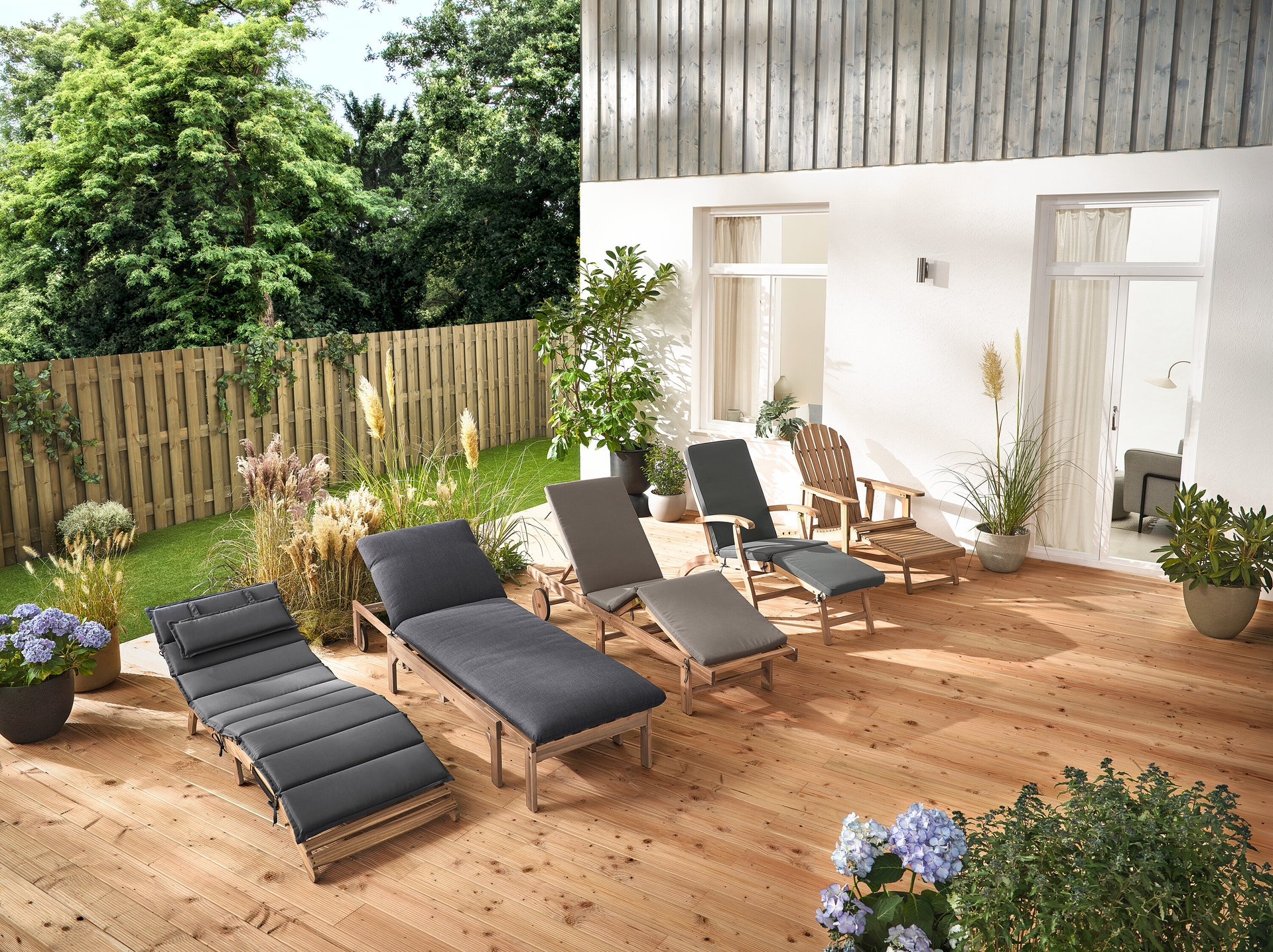 Mehrere Gartenliegen und ein Adirondack Gartensessel mit ausziehbarer Fußlehne aus Holz stehen auf einer Holzterrasse.