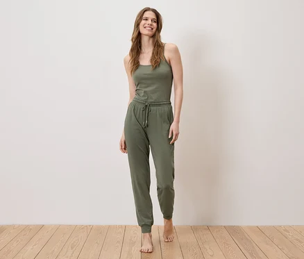 Eine Frau steht barfuß auf einem Holzboden und trägt ein olivgrünes Tanktop und eine Jogginghose.