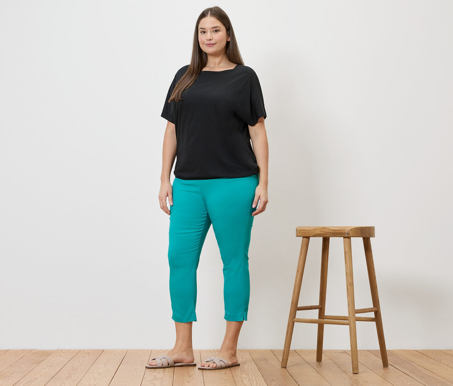 Frau in schwarzem Blusenshirt und azurblauer 7/8-Stretchhose neben einem Hocker.