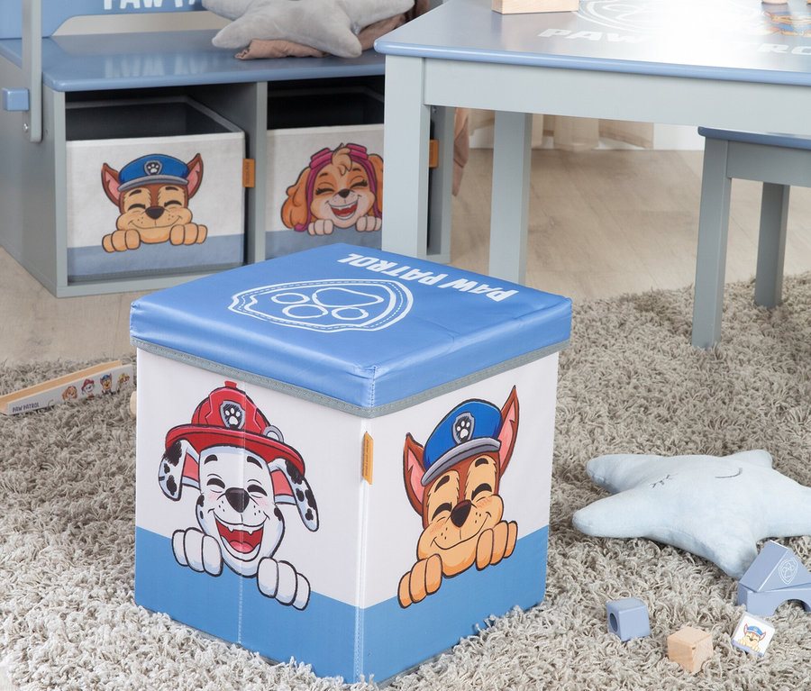 Eine blaue Paw Patrol Spielzeugkiste mit den Charakteren Marshall und Chase steht auf einem Teppich, mit einem Regal und Tisch im Hintergrund.