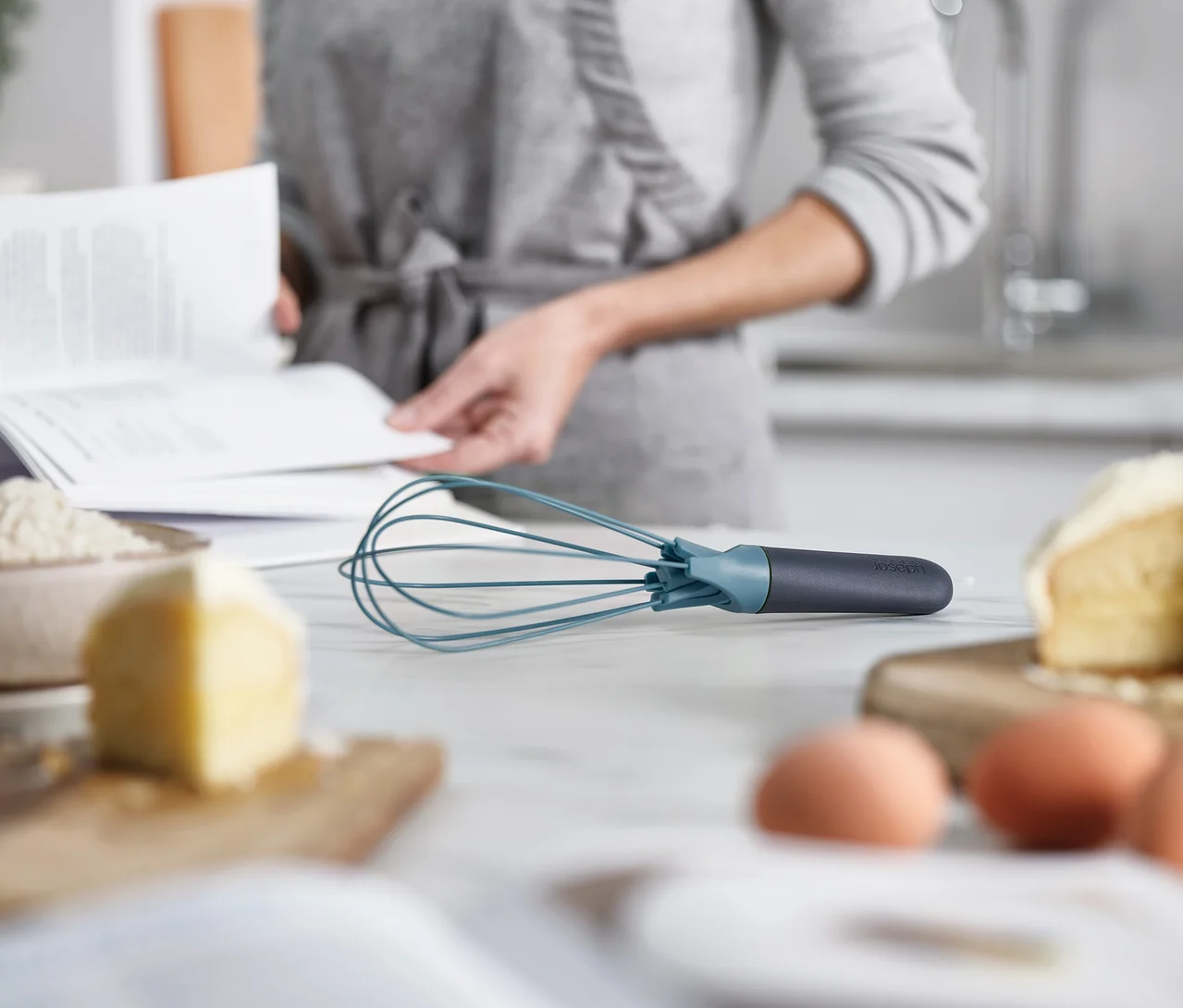 Eine Person liest ein Rezeptbuch in der Küche. Im Vordergrund liegen Eier, Butter und ein blauer Schneebesen auf einer Küchentheke.