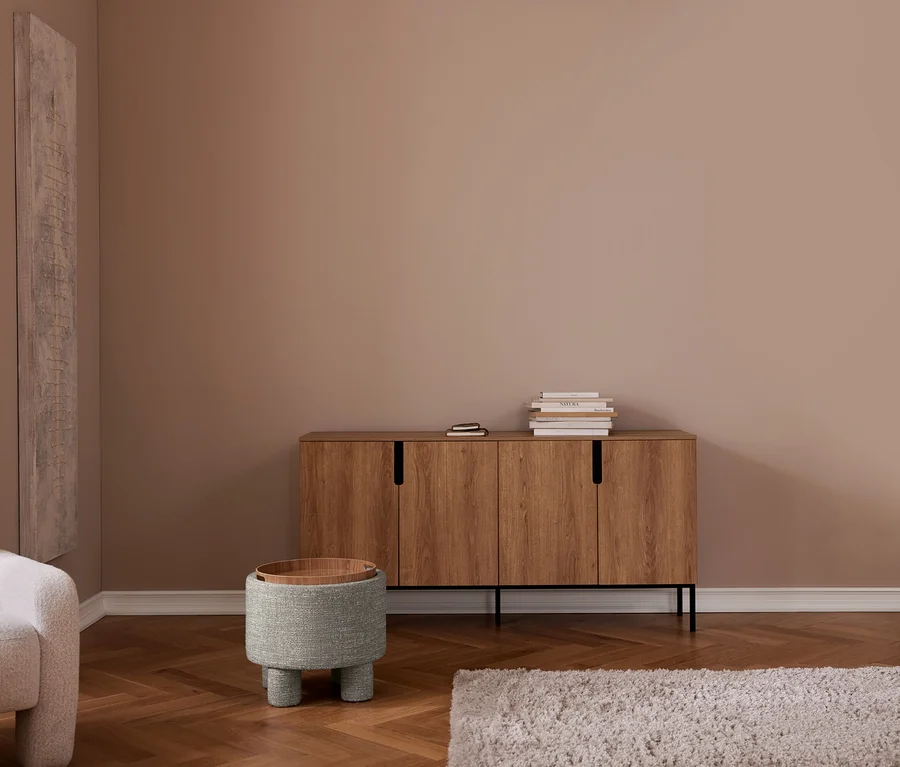 Modernes Wohnzimmer mit beigefarbenen Wänden, Fischgrätparkett. Holz-Sideboard mit Büchern, grauer Polsterhocker mit Holztablett, grauer Sessel, heller Teppich.