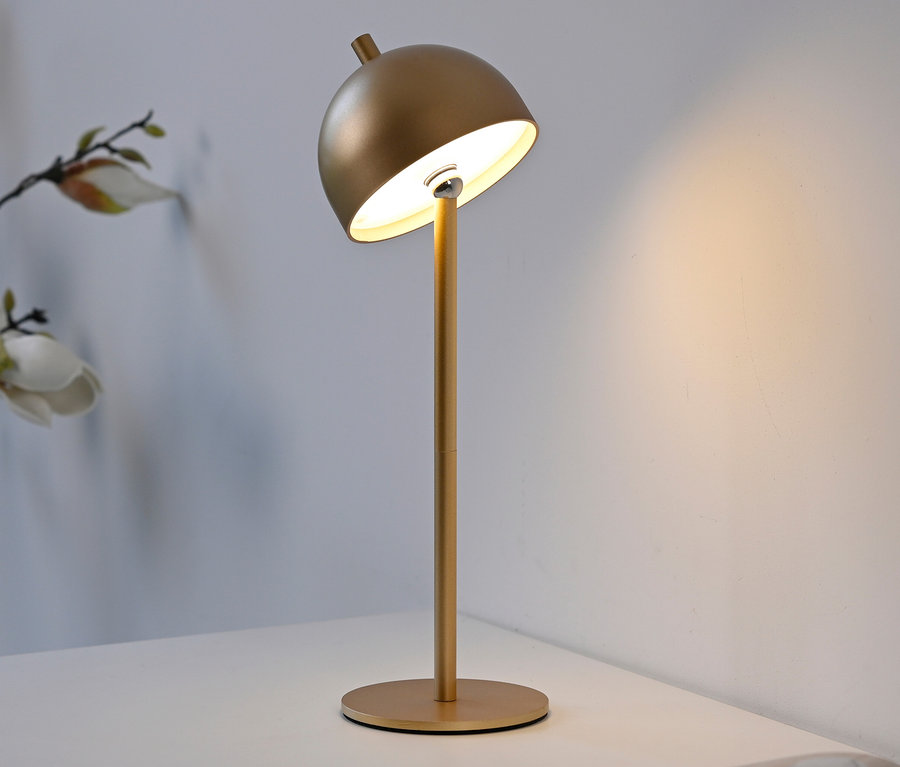 Goldfarbene Akku-Tischleuchte »Bellis« mit kuppelförmigem Schirm auf einem weißen Tisch, die warmes Licht an eine weiße Wand wirft. Im Hintergrund eine unscharfe Blumenranke.