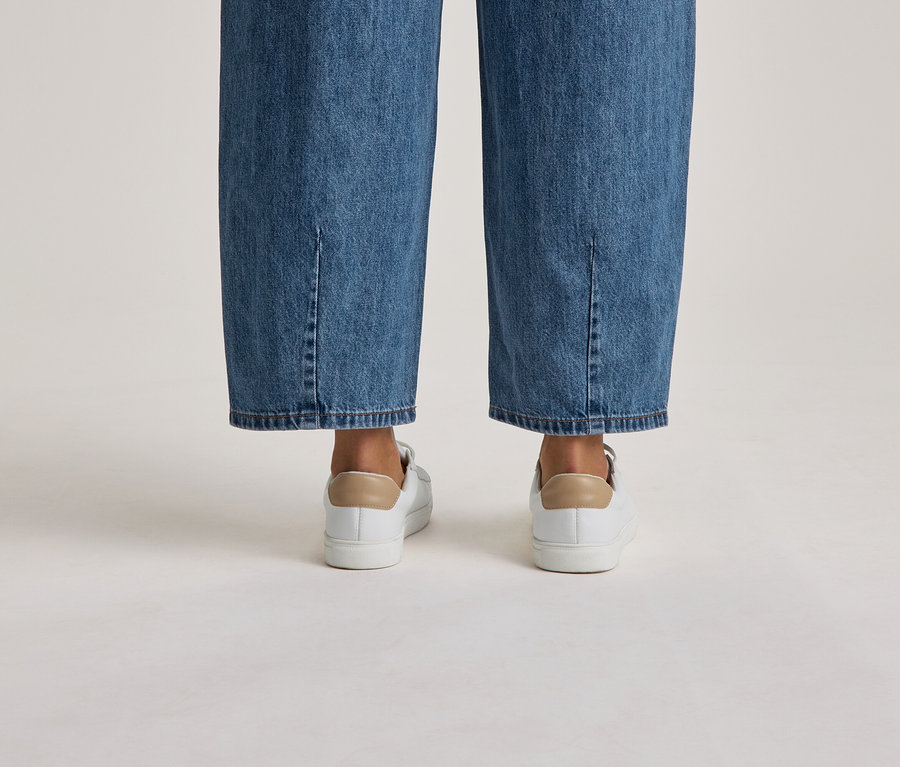 Unterschenkel einer Person in blauen Jeans mit Knöchelschlitzen und weißen Sneakern mit beigefarbenen Fersenlaschen, von hinten gesehen.