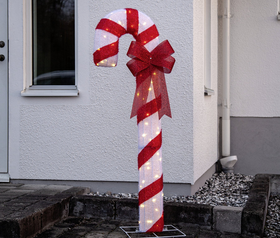 Große, beleuchtete Zuckerstange in Rot-Weiß mit roter Schleife, steht draußen vor einer weißen Hauswand auf Pflastersteinen.