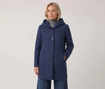 Frau in einem blauen Kapuzenmantel über einem grauen Rollkragenpullover, mit der linken Hand in der Manteltasche, blickt nach vorne.