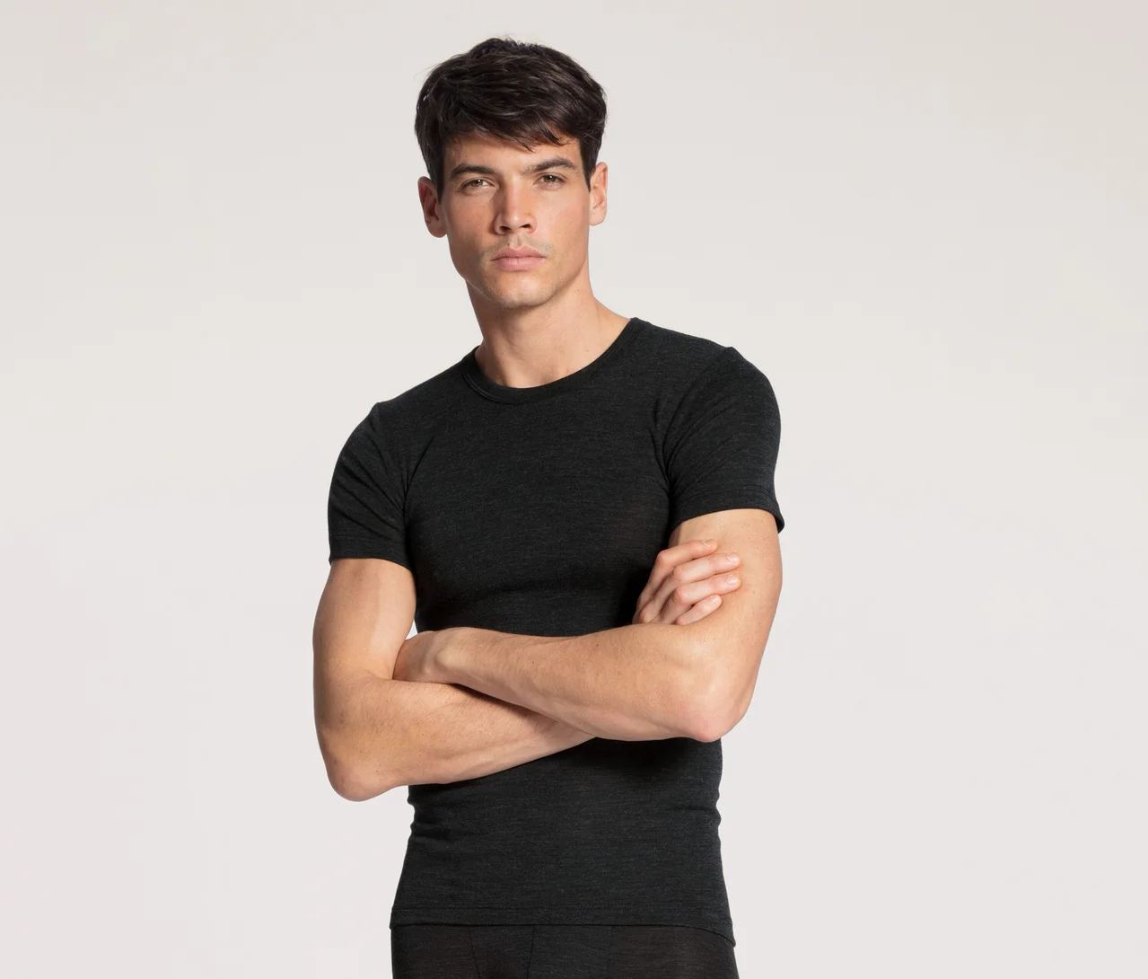 Ein Mann mit dunklem Haar und neutralem Ausdruck steht frontal da, trägt ein dunkelgraues Kurzarm-T-Shirt und dunkle Unterwäsche/Hose. Seine Arme sind vor der Brust verschränkt.