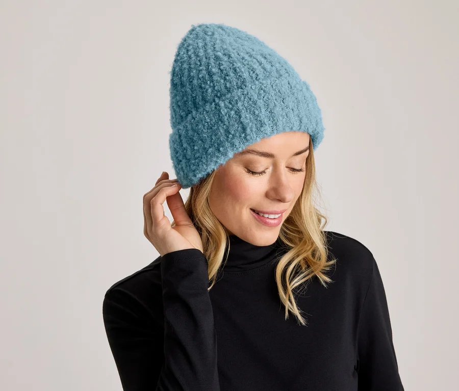 Eine lächelnde Frau mit blonden Haaren trägt eine blaue Strickmütze und einen schwarzen Rollkragenpullover. Sie blickt nach unten und hält eine Hand an ihr Ohr.