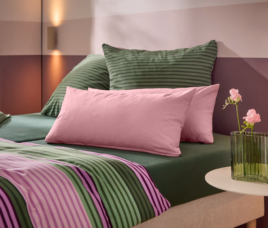 Ein Bett mit grünem Spannbetttuch, grün gestreiften Kissen, rosa Kissen und einer gestreiften Bettdecke. Ein weißer Beistelltisch trägt eine grüne gerippte Vase mit rosa Blumen. Die Wand dahinter hat horizontale rosa und lila Streifen mit zwei Wandleuchten.