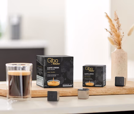 Auf einem Tisch stehen Qbo Caffè BABA BUDAN Kapseln, eine Tasse Kaffee und Kapsel-Prototypen auf einem Holzbrett.