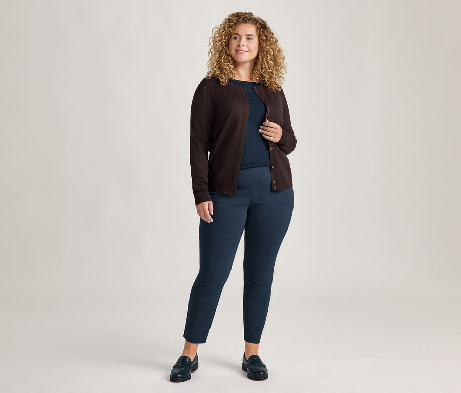 Eine lächelnde Frau mit lockigem Haar steht, trägt eine dunkelbraune Strickjacke über einem dunkelblauen Shirt mit 3/4-Arm und eine dunkelblaue Stretchhose.