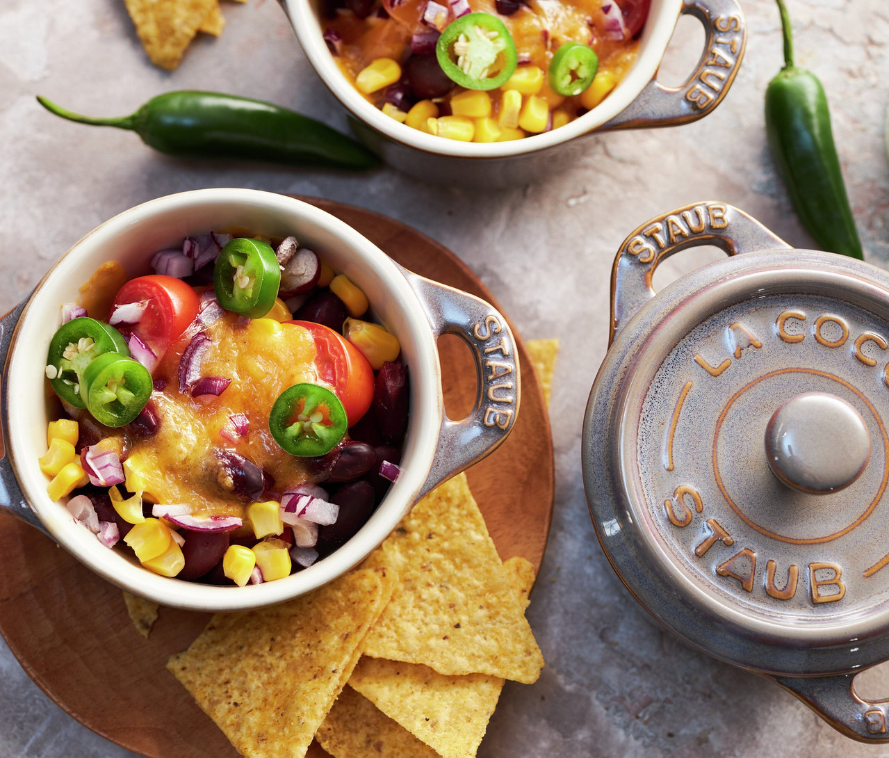 Zwei antikgraue STAUB Keramik-Cocottes mit Chili, Käse, Mais, Bohnen, Tomaten, Zwiebeln und Jalapeños, umgeben von Tortilla-Chips und ganzen Jalapeños. Ein passender Deckel ist daneben.