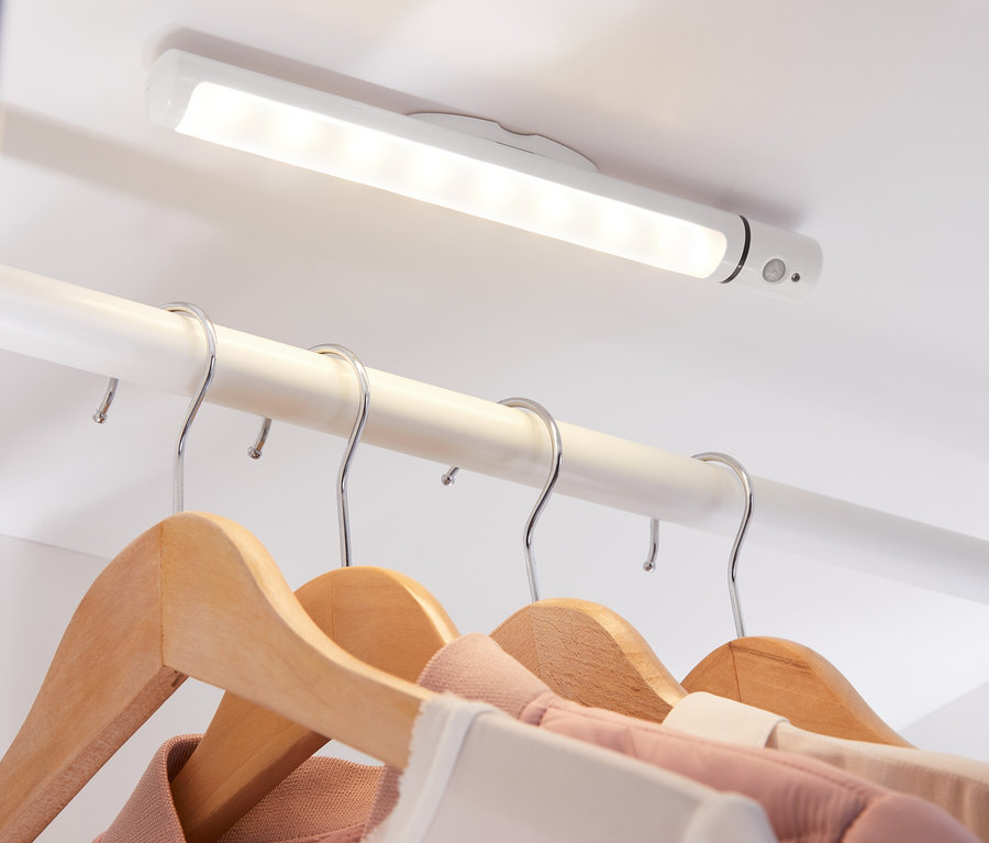 Eine LED-Multifunktionsleuchte ist an der Decke eines Schranks befestigt und beleuchtet Kleidung, die an Holzbügeln hängt.