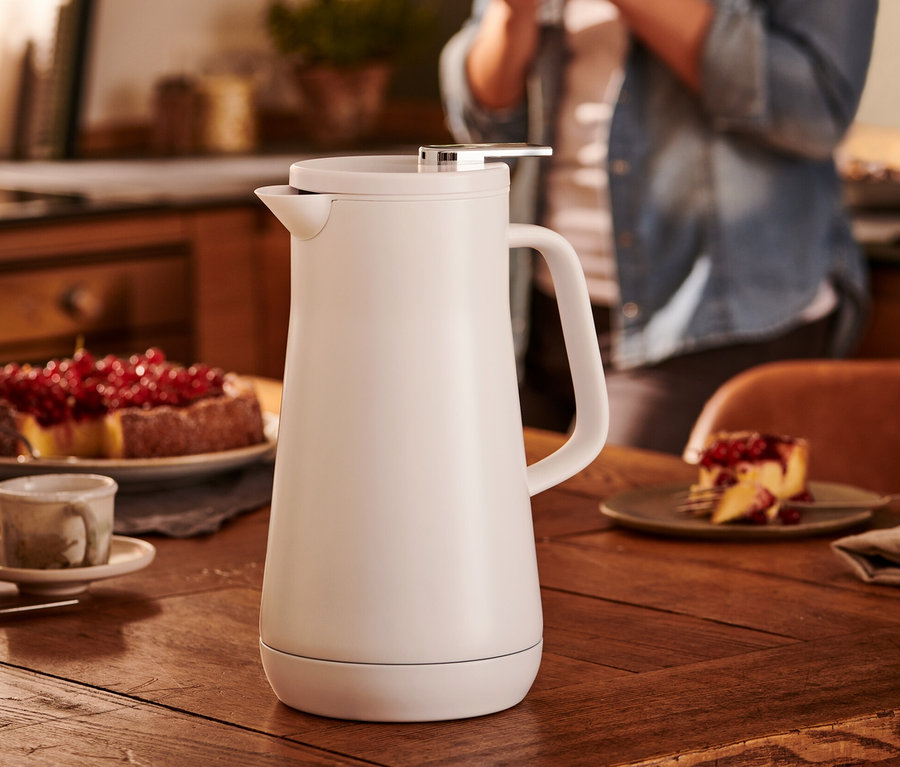 Auf einem Holztisch steht eine weiße Isolierkanne, 1l. Im Hintergrund hält eine Frau eine Tasse.