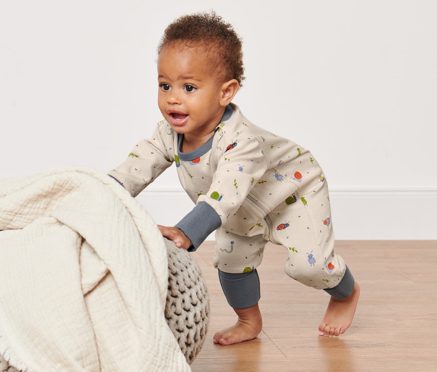Krabbelndes Kind im Baby-Pyjama mit Reißverschluss, das sich an einem Pouf mit Decke abstützt.