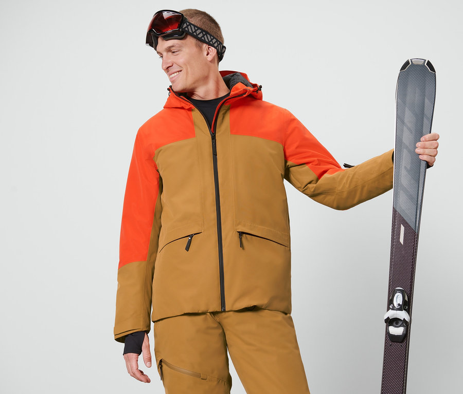 Mann mit Skibrille hält Ski und trägt eine orange-braune Skijacke.