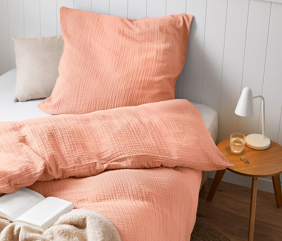 Musselin-Bettwäsche in Normal- und Übergröße auf einem Bett mit Buch und Lampe.