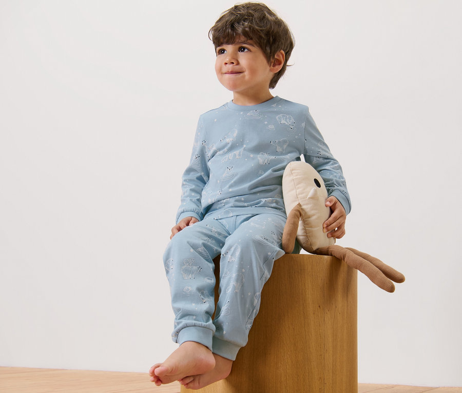Ein Junge sitzt auf einem Hocker und hält ein Stofftier. Er trägt einen blauen Kinder-Pyjama (2 Stück).