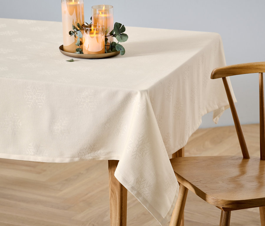 Eine normal große, cremefarbene Jacquard-Tischdecke liegt auf einem Tisch neben einem Holzstuhl. Auf der Tischdecke stehen Kerzen.