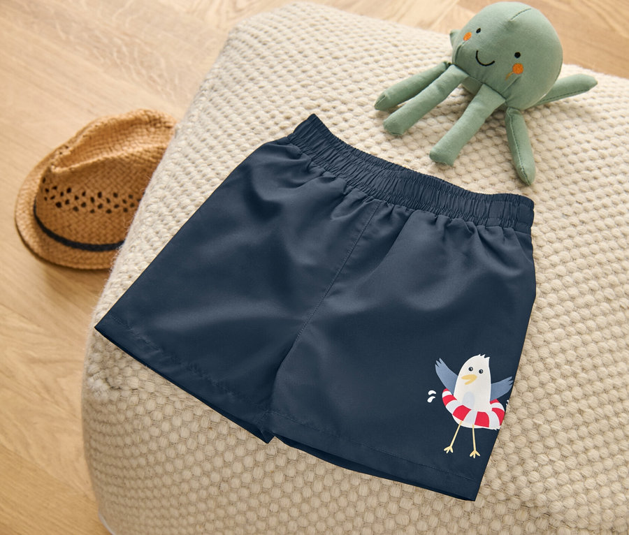 Kinder-Boardshorts, maritim, liegen auf einem Pouf. Daneben liegen ein Stofftier-Oktopus und ein Strohhut.