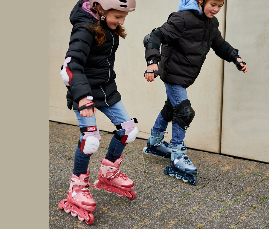 Zwei Kinder fahren mit HUDORA Inline Skates »Mia 2.0« mit Schutzausrüstung und Helmen.