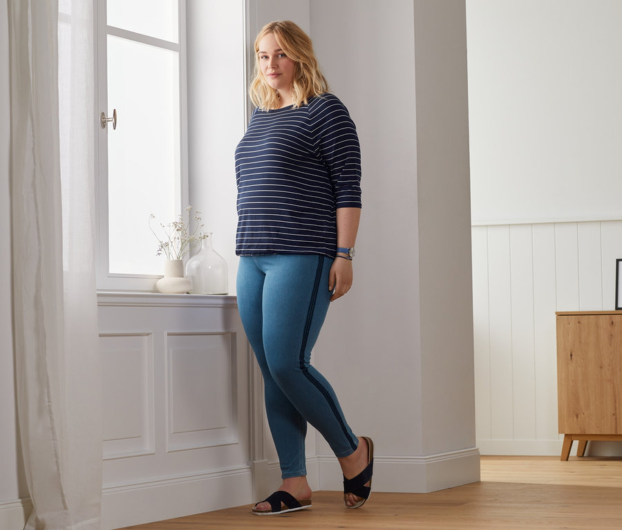 Blonde Frau posiert in einem Shirt mit Fledermausärmeln und Jeggings am Fenster.