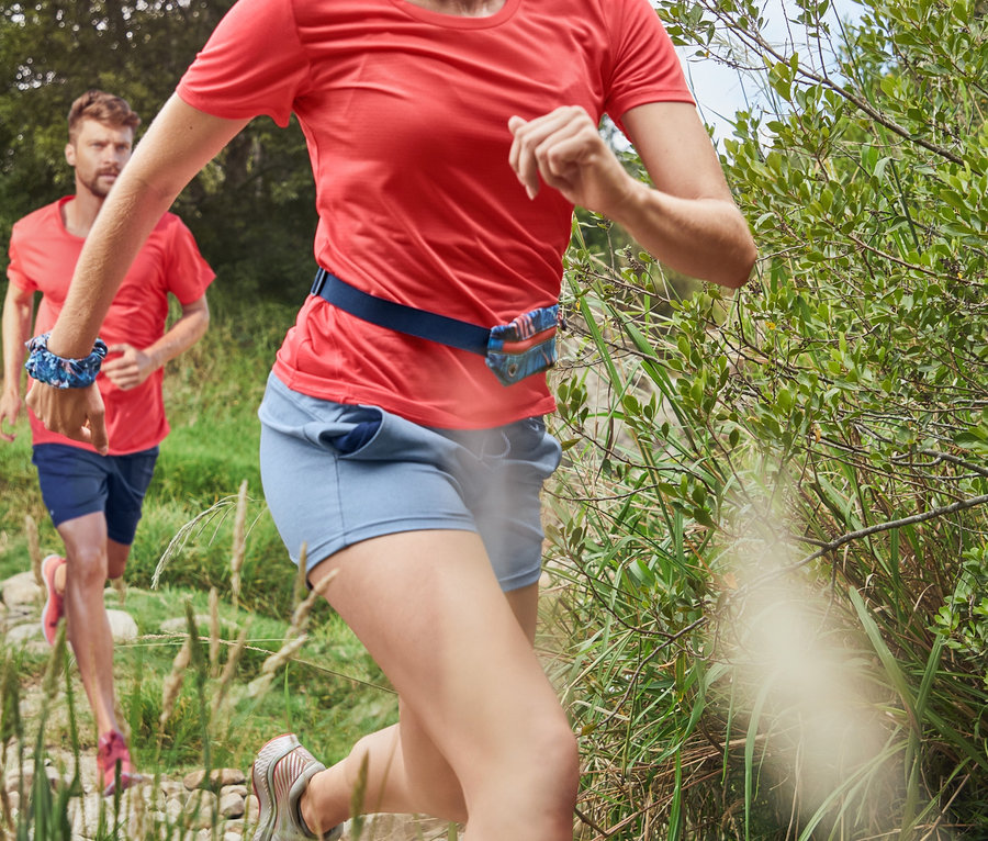 Eine Frau in einem korallefarbenen Funktionsshirt und einer dunkelblauen 3/4-Sporttight läuft auf einem steinigen Pfad. Im Hintergrund läuft ein Mann in Funktionsshorts und einem korallefarbenen Sportshirt.